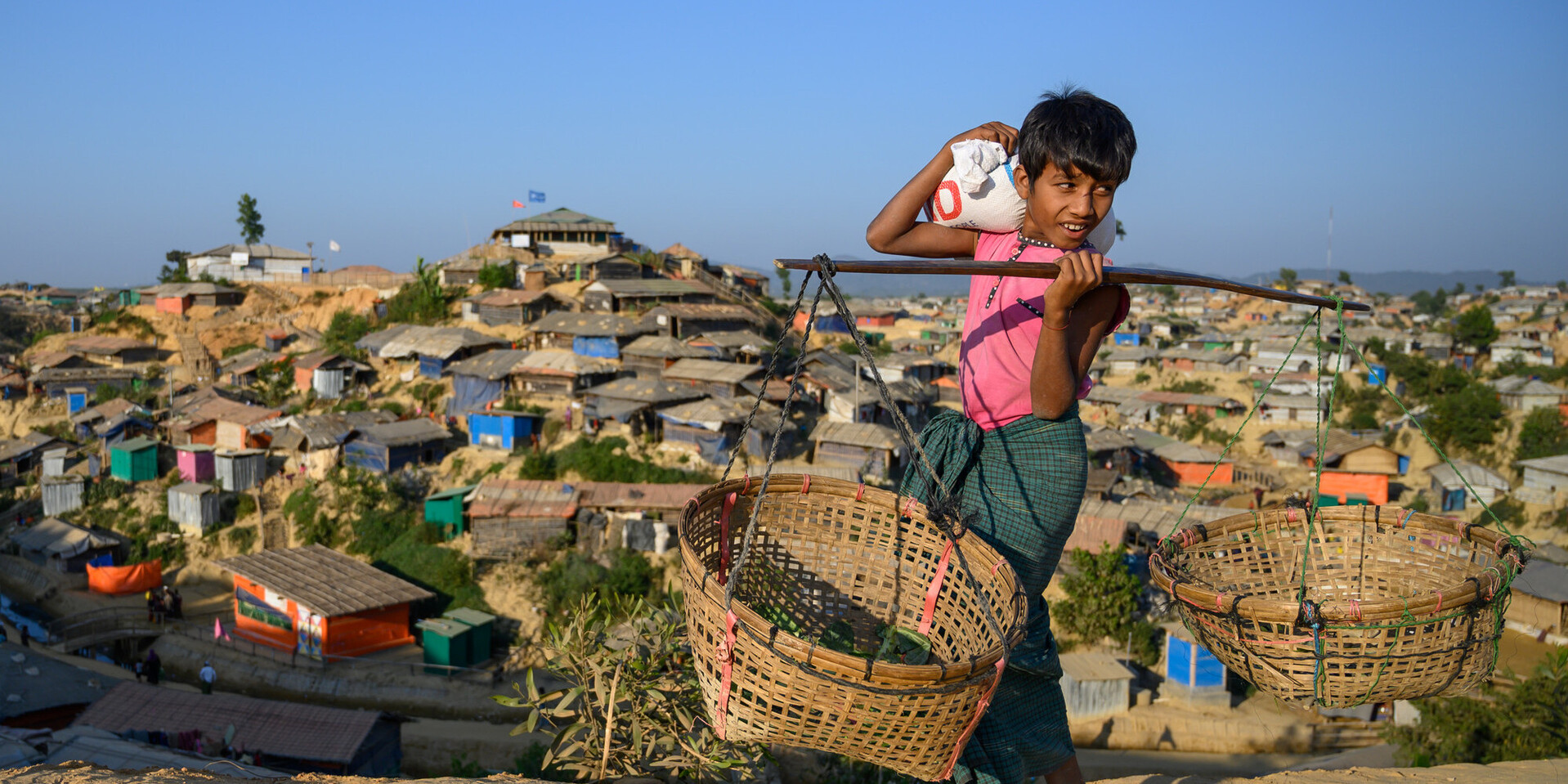 Ein Kind steht mit zwei Körben vor dem Flüchtlingscamp in Cox’s Bazar, Bangladesch