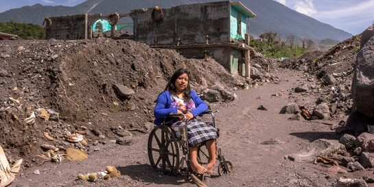 Eine Frau in Guatemala sitzt in einem Rollstuhl. Im Hintergrund der Vulkan Fuego