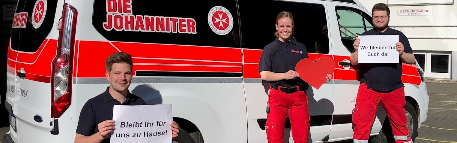 Drei Mitarbeiter der Johanniter-Unfall-Hilfe halten vor einem Einsatzwagen Schilder in den Händen