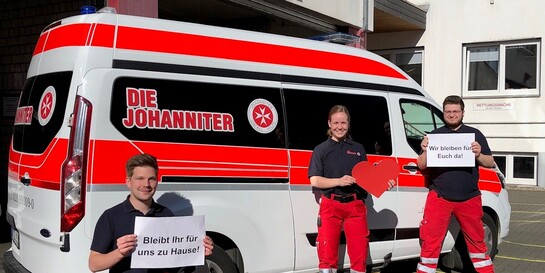 Drei Mitarbeiter der Johanniter-Unfall-Hilfe halten vor einem Einsatzwagen Schilder in den Händen