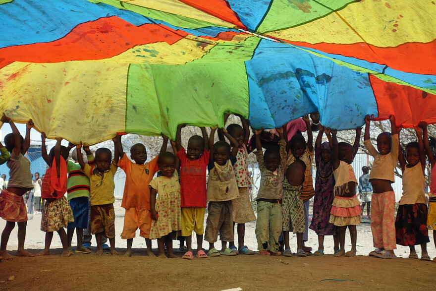 Kinder im Geflüchtetencamp Kakuma spielen mit einem bunten Schwungtuch Kinder im Geflüchtetencamp Kakuma spielen mit einem bunten Schwungtuch