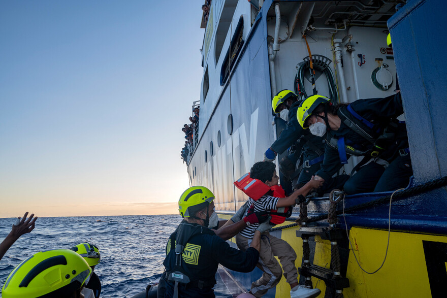 Eine Gruppe von Seenotretter:innen heben ein geflüchtetes Kind in ein Boot