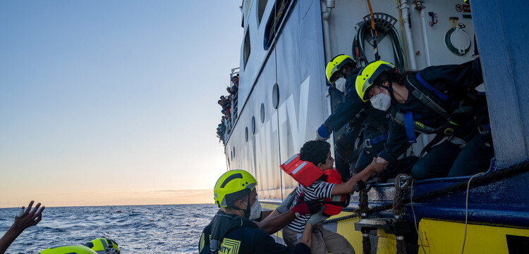 Eine Gruppe von Seenotretter:innen heben ein geflüchtetes Kind in ein Boot Eine Gruppe von Seenotretter:innen heben ein geflüchtetes Kind in ein Boot