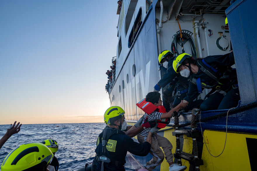 Eine Gruppe von Seenotretter:innen heben ein geflüchtetes Kind in ein Boot Eine Gruppe von Seenotretter:innen heben ein geflüchtetes Kind in ein Boot