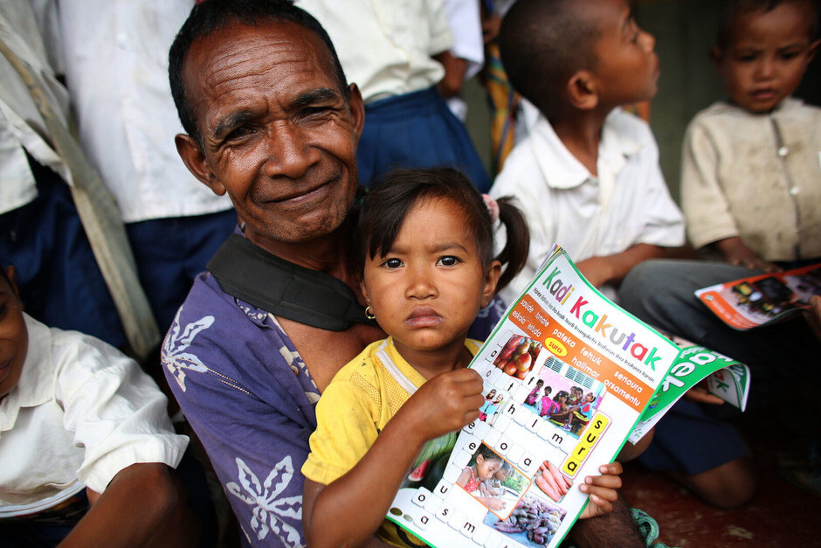 Auf dem Schoß eines Mannes in Laos sitzt ein Mädchen mit Kinderzeitschrift in den Händen