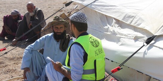 Vier Männer, einer davon ist aufgrund einer Warnweste als humanitärer Helfer erkennbar, sitzen vor einer Notunterkunft in Afghanistan, zwei Männer unterhalten sich