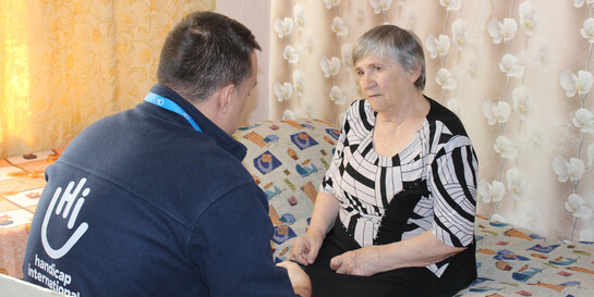 Eine ältere Frau sitzt auf einem Bett und schaut einen Mann an, der vor ihr sitzt. Er trägt die Aufschrift Handicap International auf seinem Rücken