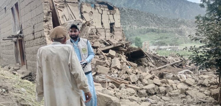Ein Helfer steht bei einem Mann in Afghanistan, neben ihnen ist ein Haus, das nach einem Erdbeben zum Teil eingestürzt ist