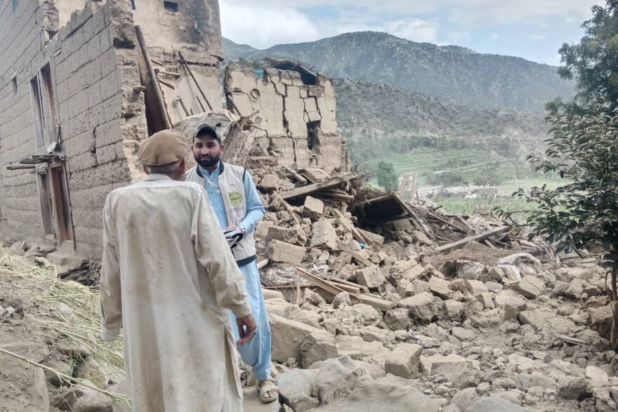 Ein Helfer steht bei einem Mann in Afghanistan, neben ihnen ist ein Haus, das nach einem Erdbeben zum Teil eingestürzt ist