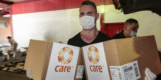 Eine Helfer mit Mundschutz und Handschuhen trägt zwei CARE-Pakete