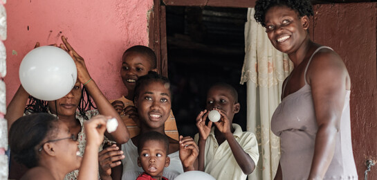 Eine Frau steht mit Liftballons spielenden Kindern vor einem Haus in Haiti.