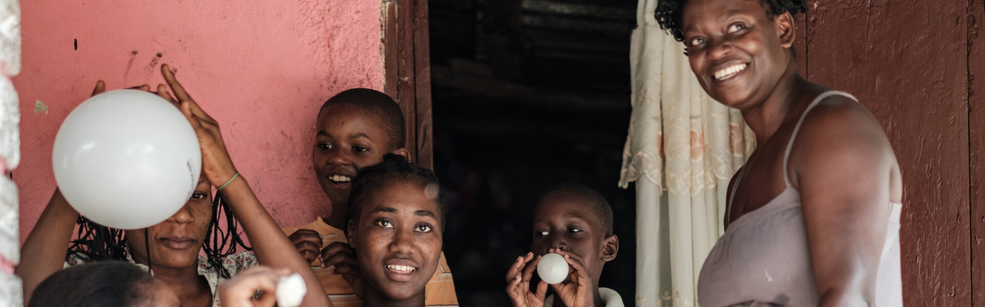 Eine Frau steht mit Liftballons spielenden Kindern vor einem Haus in Haiti.