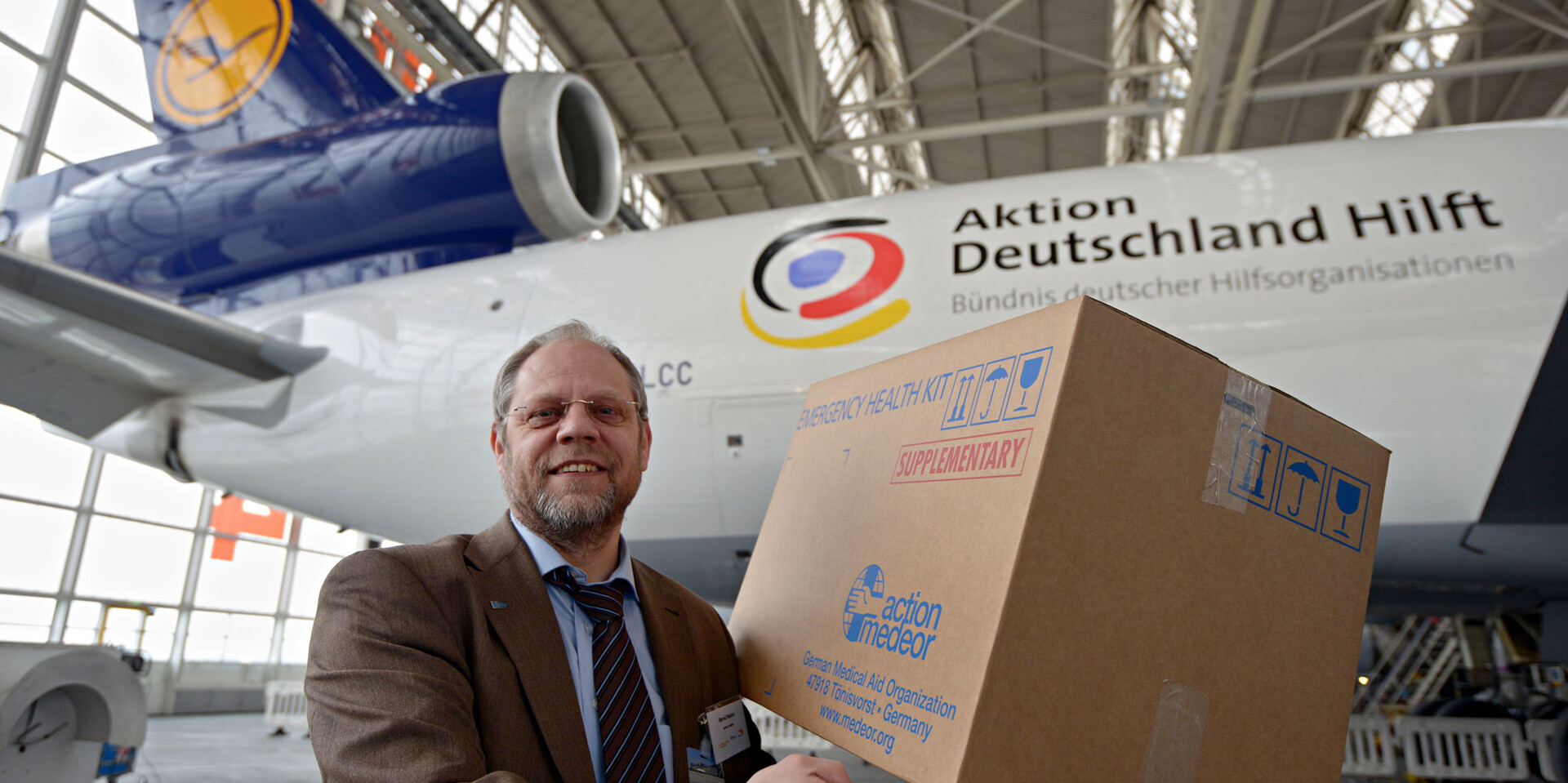 Ein Mitarbeiter aus dem Bündnis steht vor einem Flugzeug und hält ein Hilfspaket in den Händen