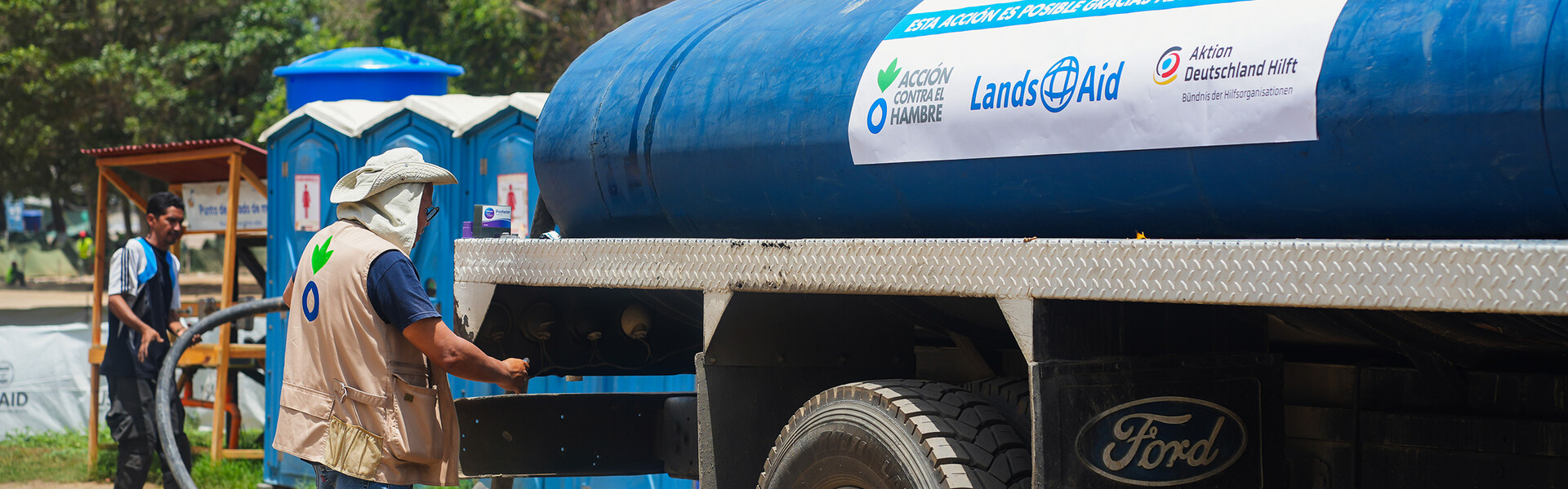 Ein Helfer steht neben einem Wassertank mit der Aufschrift Aktion gegen den Hunger, LandsAid, Aktion Deutschland Hilft