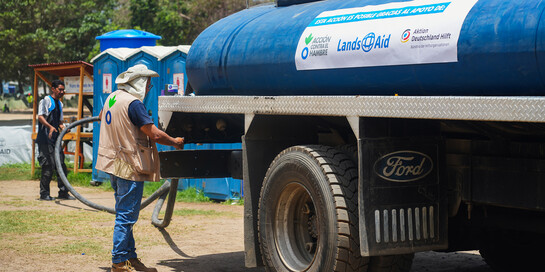Ein Helfer steht neben einem Wassertank mit der Aufschrift Aktion gegen den Hunger, LandsAid, Aktion Deutschland Hilft