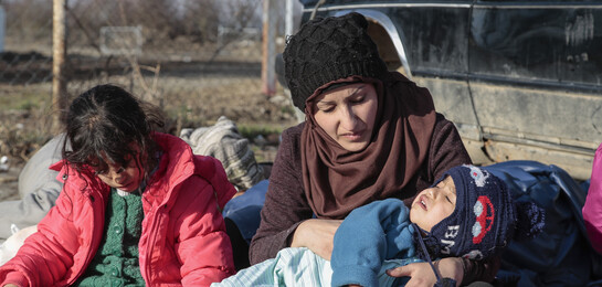 In Syrien hält eine Frau ein Kind neben einem Auto. Es ist eine Flüchtlingsfamilie.