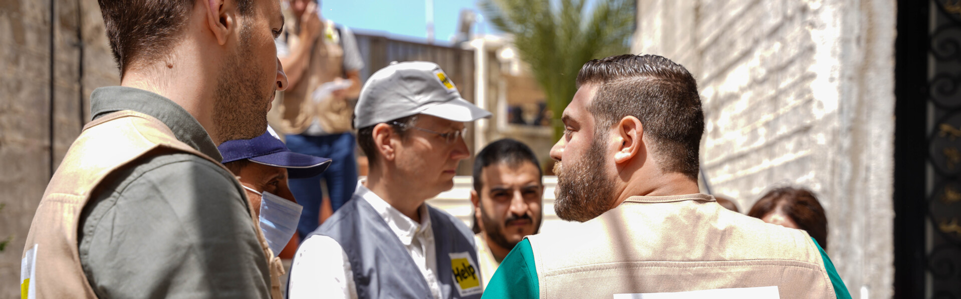 Mehrere Männer stehen neben Gebäuden in Syrien, sie tragen die Aufschrift Help Hilfe zur Selbsthilfe auf ihren Westen