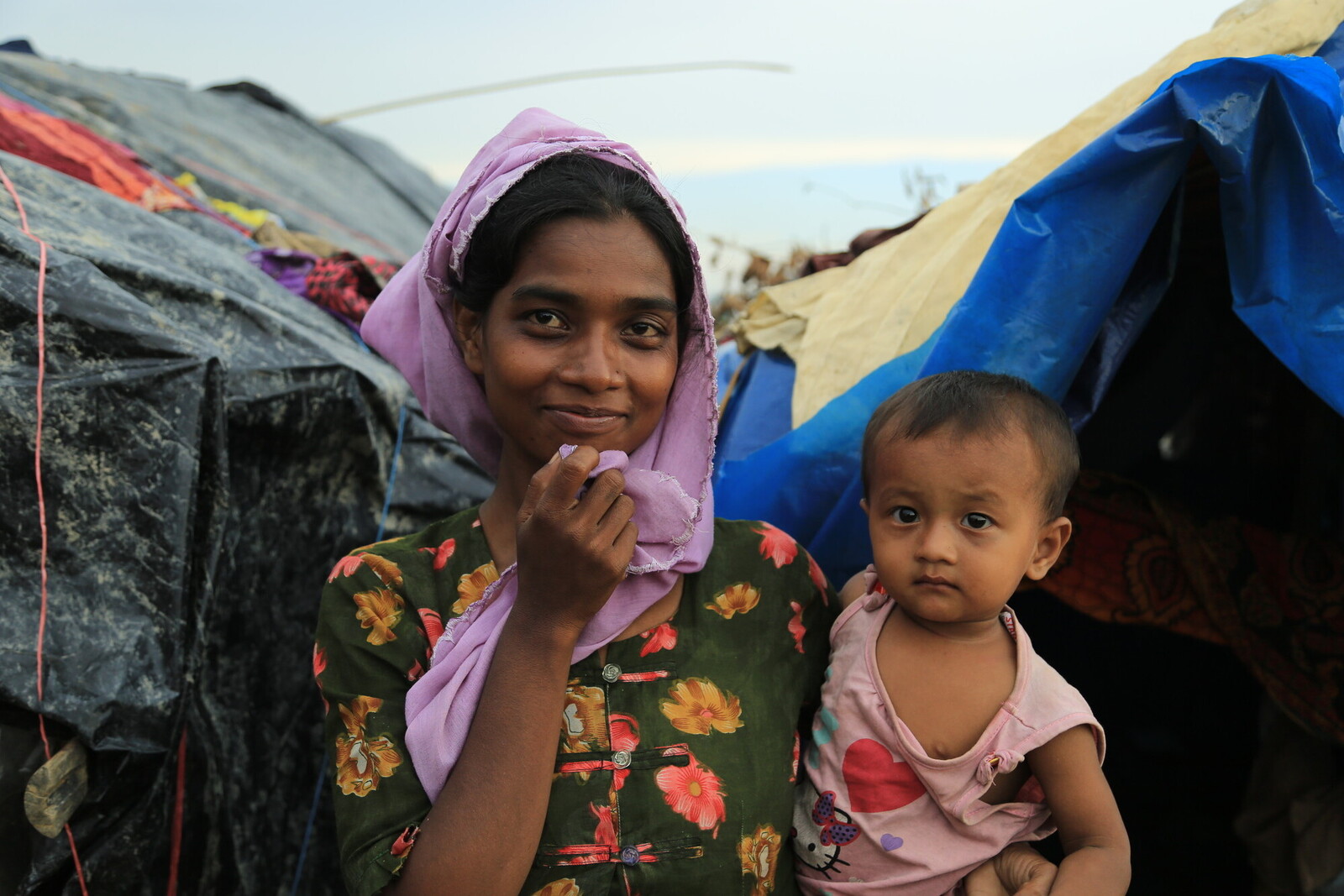 Die Frau in Bangladesch hält ihr kleines Kind im Arm und lächelt zuversichtlich vor ihrer Notunterkunft