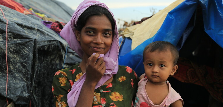 Die Frau in Bangladesch hält ihr kleines Kind im Arm und lächelt zuversichtlich vor ihrer Notunterkunft Die Frau in Bangladesch hält ihr kleines Kind im Arm und lächelt zuversichtlich vor ihrer Notunterkunft
