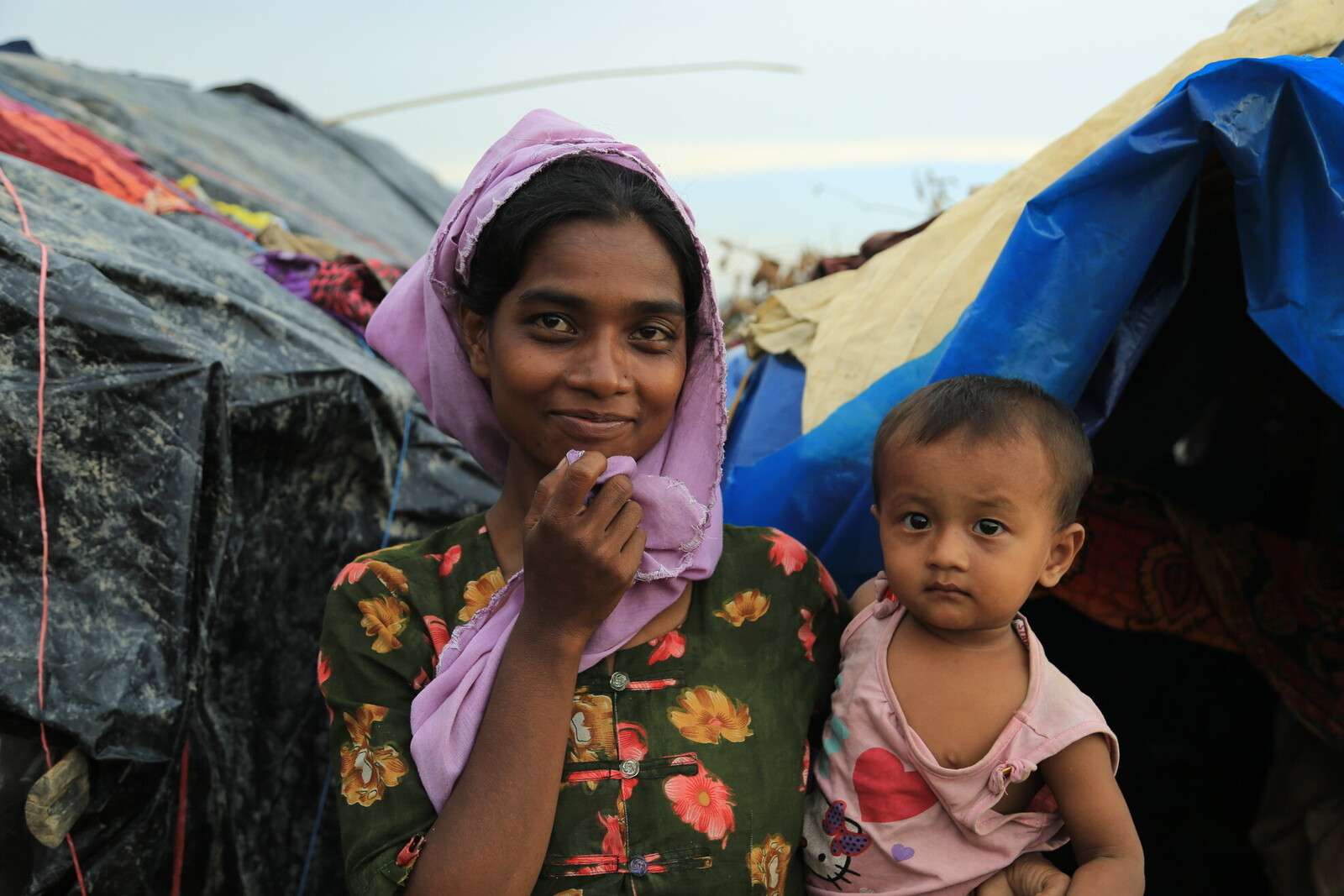 Die Frau in Bangladesch hält ihr kleines Kind im Arm und lächelt zuversichtlich vor ihrer Notunterkunft