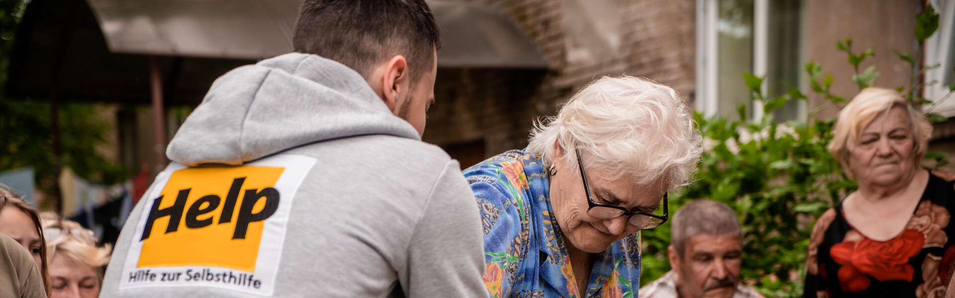 Ein Helfer von der Organisation Help – Hilfe zur Selbsthilfe überreicht einer Frau einen Beutel mit Hilfsgütern