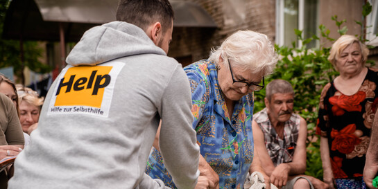 Ein Helfer von der Organisation Help – Hilfe zur Selbsthilfe überreicht einer Frau einen Beutel mit Hilfsgütern