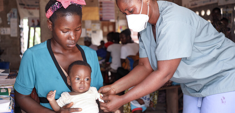 Ein Baby wird in Sierra Leone auf dem Schoß seiner Mutter von einer Helferin untersucht