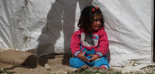 In Syrien sitzt ein Flüchtlingskind vor einem Vorhang.