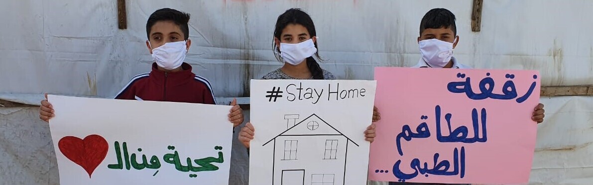 Drei syrische Kinder halten Schilder hoch: Sie fordern, während der Coronakrise zu Hause zu bleiben