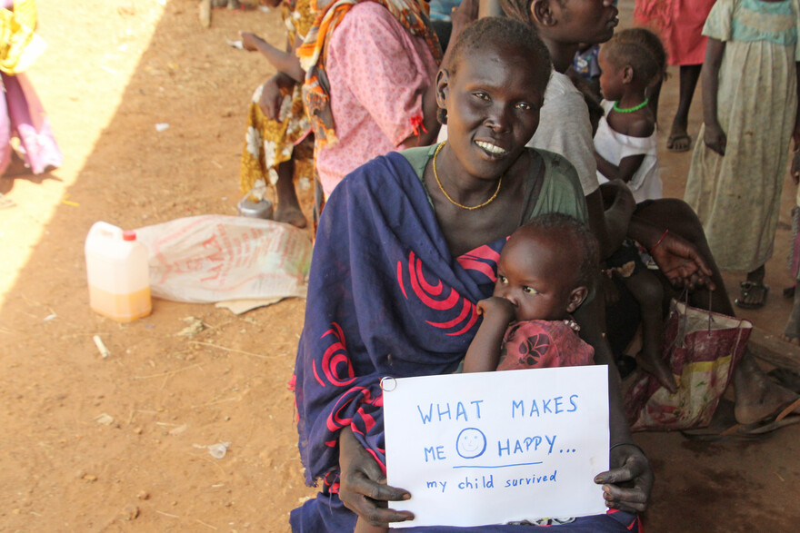 Eine Frau hält ihr Kind im Arm und einen Zettel mit der Aufschrift "What makes me happy - my child survived"