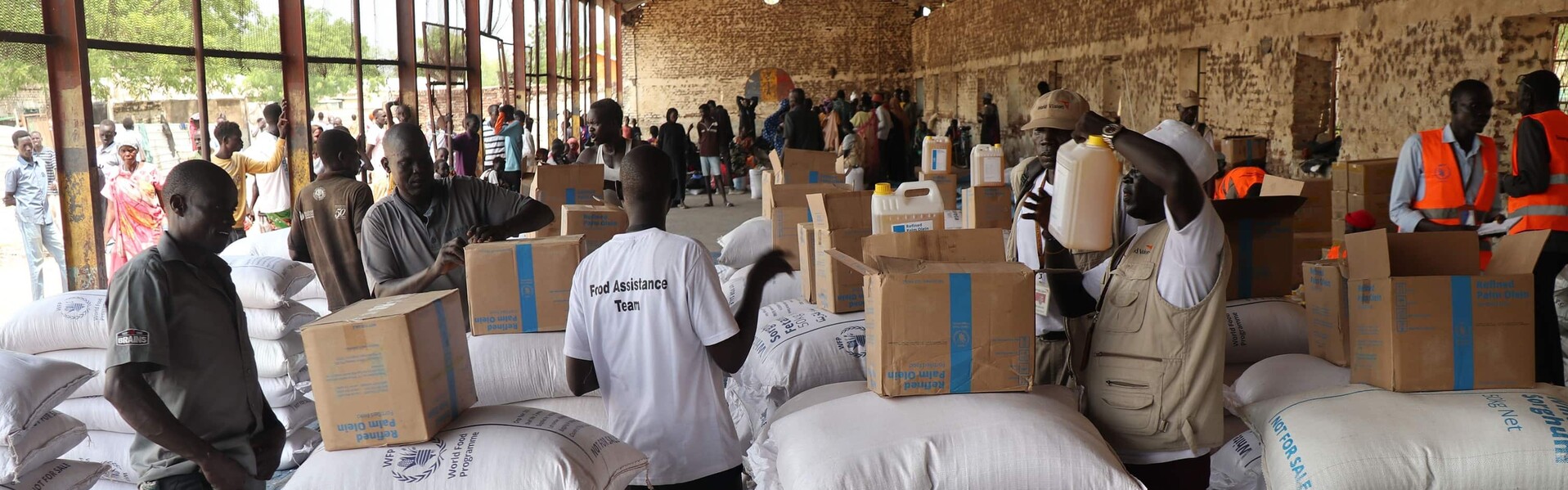 Hilfsorganisationen fordern Schutz der bevorstehenden Ernten Helfende packen Kisten in einer Halle. Im Vordergrund liegen übereinandergestapelte weiße Säcke