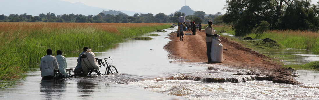 Männer in Uganda schieben ihre Fahrräder über eine überschwemmte Straße