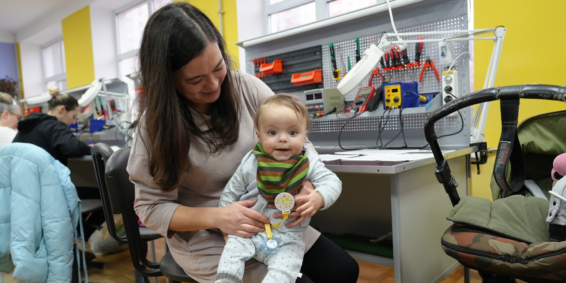 Eine Frau mit Baby auf dem Schoß sitzt vor einer Wand mit Schaltknöpfen, Kabeln und Werkzeugen. Im Hintergrund weitere Frauen, die an den Schaltknöpfen arbeiten