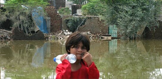 Flut Pakistan: Mädchen mit Wasserflasche steht vor überschwemmtem Gebiet