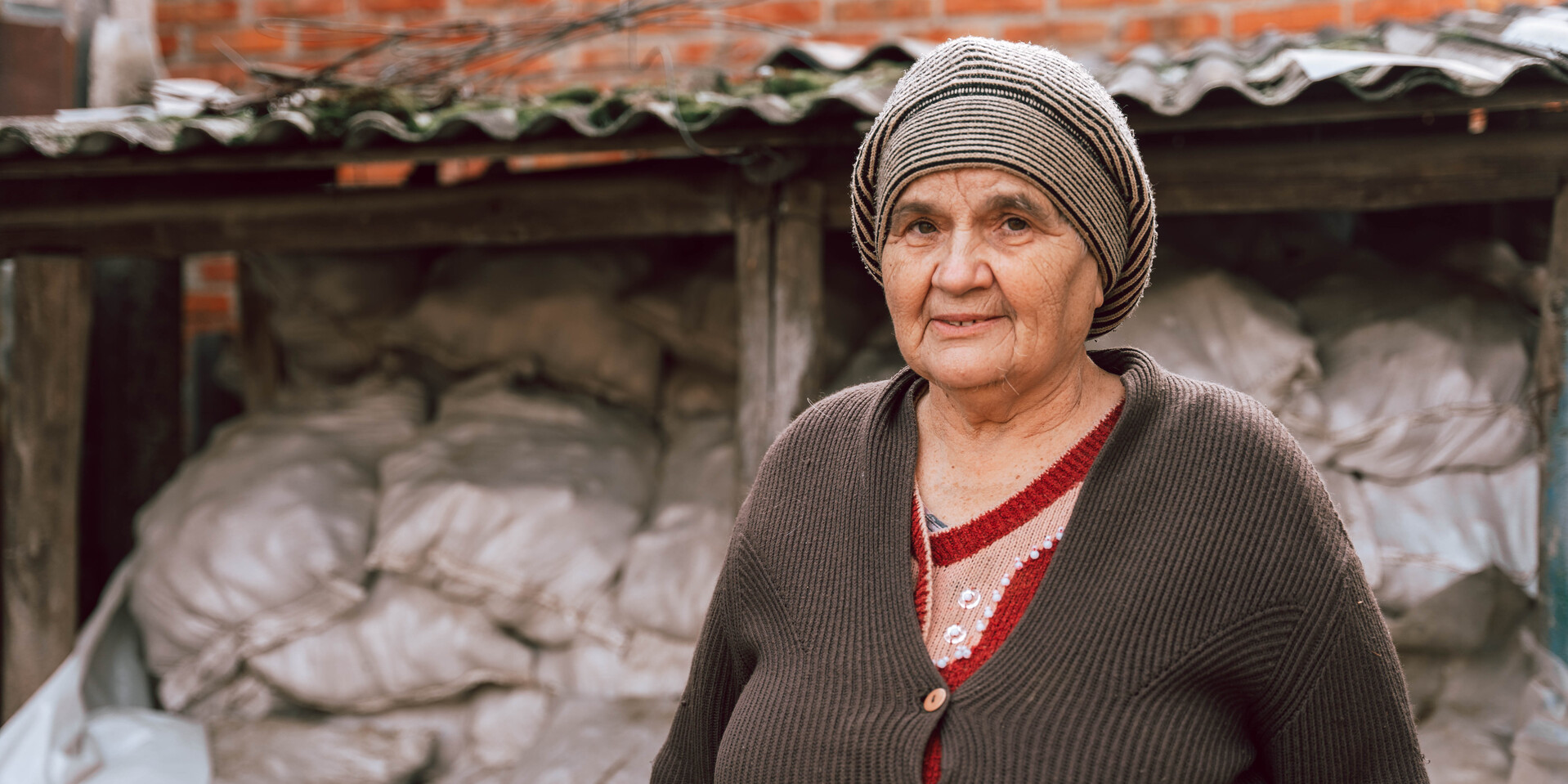 Hanna ist dankbar über die Winterhilfe durch Help Eine ältere Frau mit Mütze steht vor einem mit Holzbriketts gefüllten Schuppen neben einem Haus in der Ukraine und schaut in die Kamera