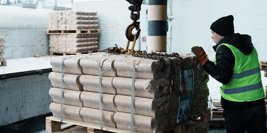 Ein Mann mit gelber Sicherheitsweste steht hinter einer mit Holzbriketts beladenen Palette