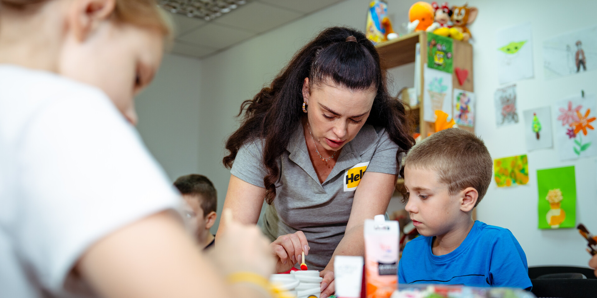 Help leistet psychosoziale Hilfe und biete Räume für Kinder, in denen sie basteln können Drei Kinder und eine Erwachsene, die die Aufschrift Help auf ihrem Oberteil trägt, basteln in einem Raum, an dessen Wände gemalte Bilder hängen