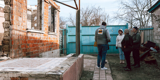Fünf Personen stehen im Bereich des Vorgartens eines zerstörten Hauses in der Ukraine. Einer der Männer trägt die Aufschrift Help auf dem Rücken