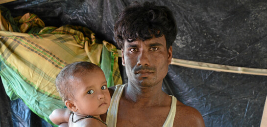 Ein Mann steht mit seinem Kind unter einem Zelt. Sie sind beide Rohingya-Flüchtlinge.